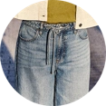 Model trägt ein weißes Leinenhemd und eine beige Leinenhose, sitzt auf einem Stuhl und blickt nach rechts