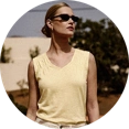 Model trägt ein weißes Leinenhemd und eine beige Leinenhose, sitzt auf einem Stuhl und blickt nach rechts