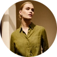  Model trägt ein weißes Leinenhemd und eine beige Leinenhose, sitzt auf einem Stuhl und blickt nach rechts