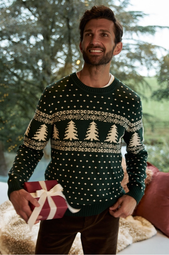 Mann trägt einen dunkelgrünen Weihnachtspullover mit Tannenbaummuster und hält ein verpacktes Geschenk