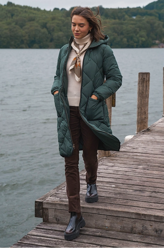 Frau trägt einen gesteppten Maximalmantel und Stiefel an einem See