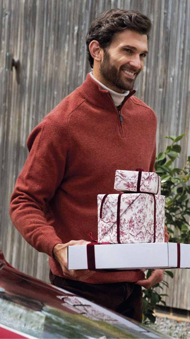 Mann trägt einen roten Pullover mit Reißverschluss und hält verpackte Weihnachtsgeschenke im Freien